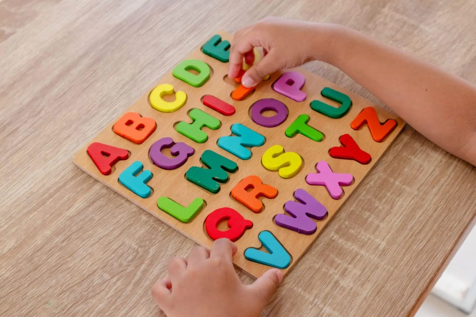 Mãos de uma criança encaixando letras coloridas num quebra-cabeça de alfabeto de madeira